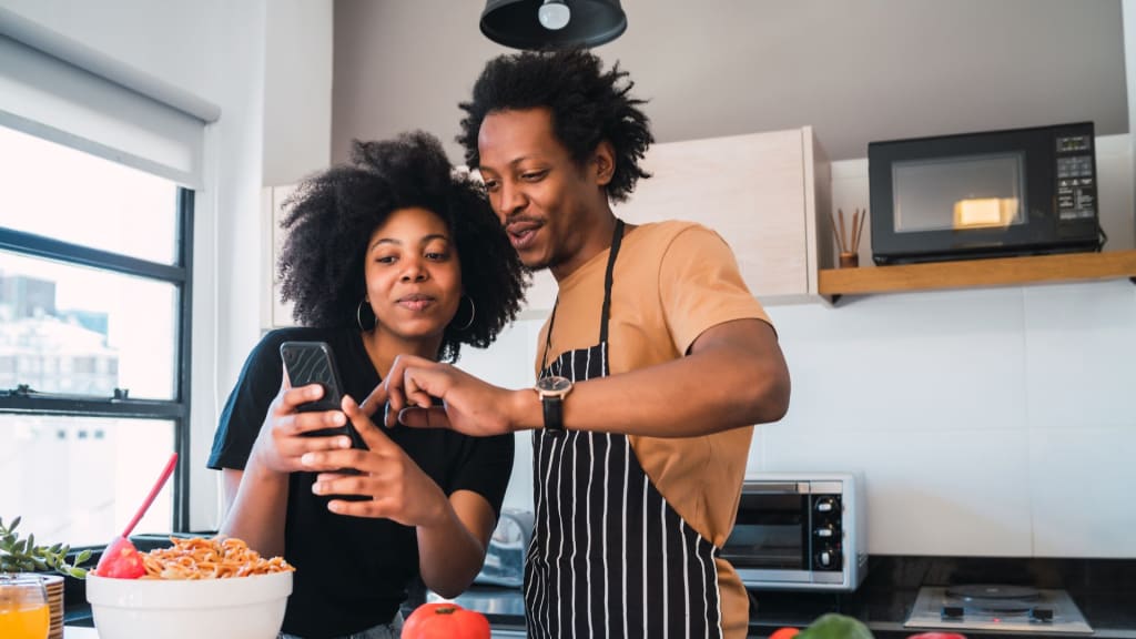 Afro-descendant couple in their 30s, using a phone in the kitchen while cooking together. The man, standing behind the woman, looks at the phone with her. The kitchen is clean and modern.